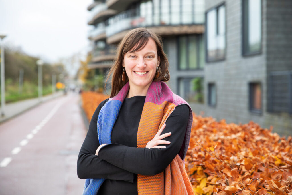 Vrouw met lange bruine haren en kleurrijke sjaal poseert lachend buiten voor een moderne woonwijk en herfstige struiken.