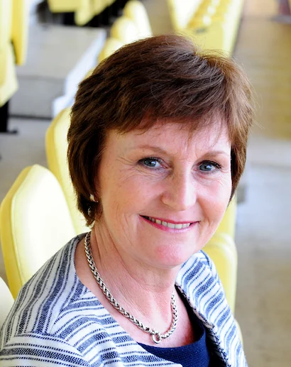 Vrouw genaamd Rita Breikers glimlacht terwijl ze op een gele stadionstoel zit.