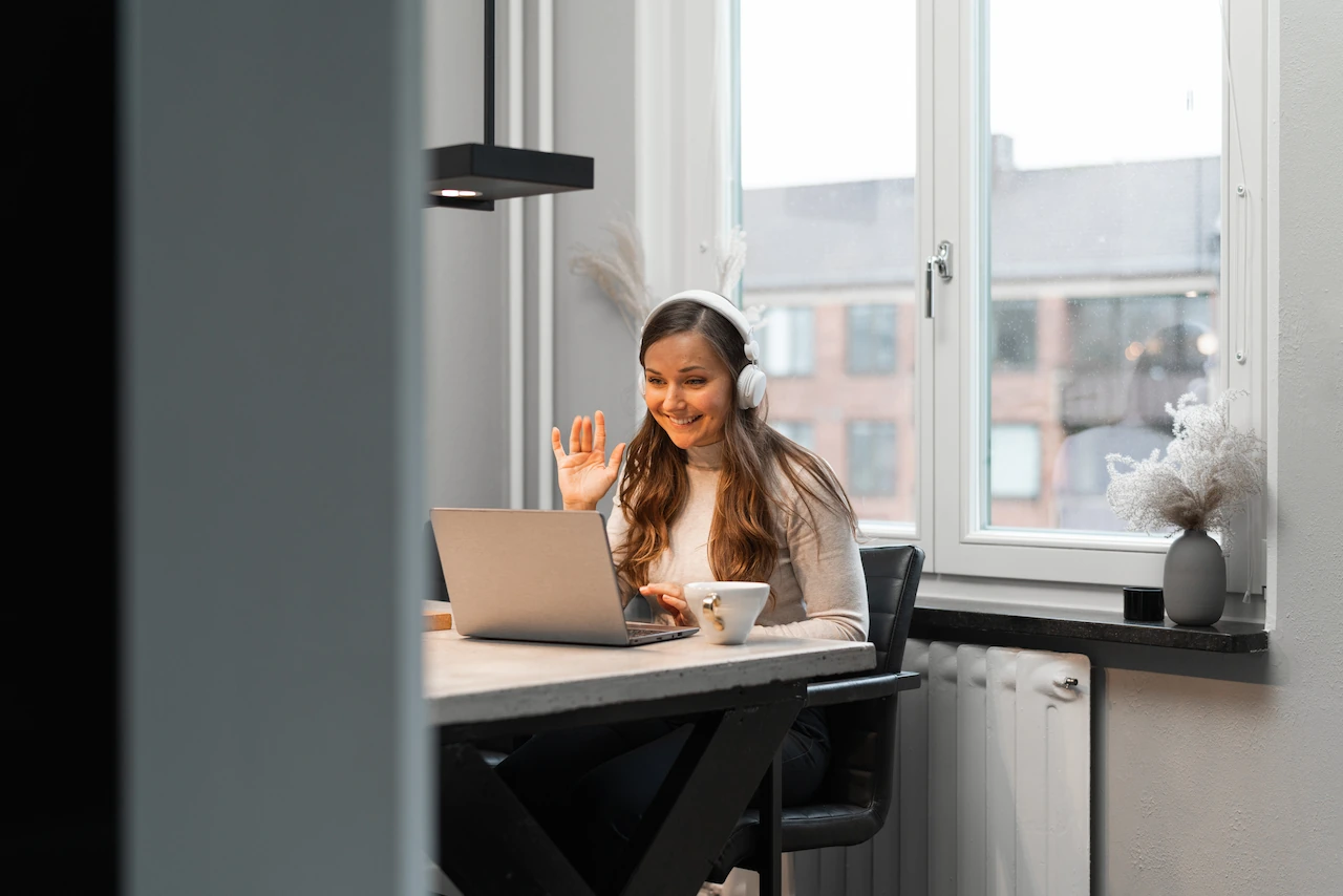 Vrouw werkt thuis aan tafel met laptop en draagt een koptelefoon tijdens een videogesprek.
