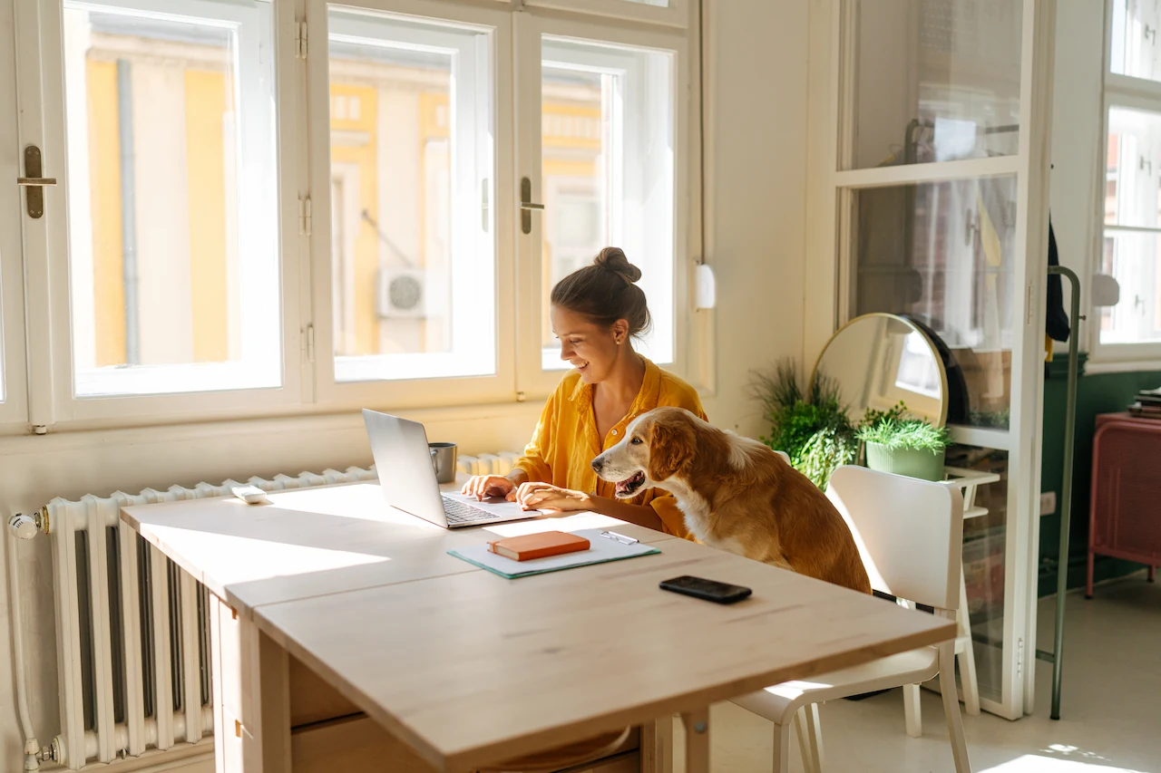 Vrouw werkt thuis aan een laptop met een hond naast zich aan tafel.