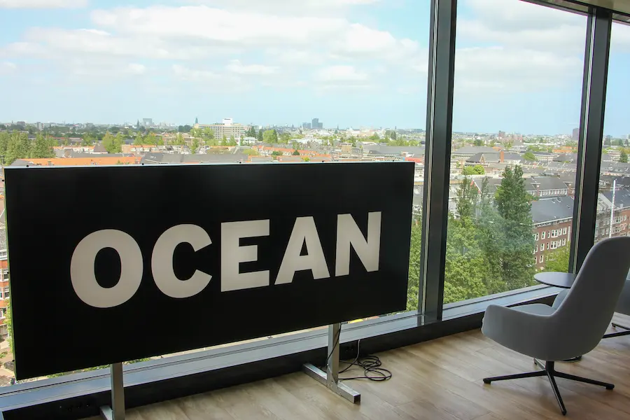 Zwart bord met het woord "OCEAN" in witte letters voor een groot raam met uitzicht over een stad.