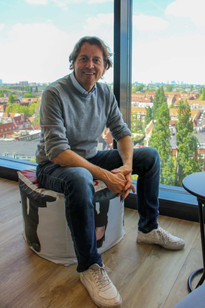 Man in grijze trui zit glimlachend op een poef voor een groot raam met uitzicht over een stad.