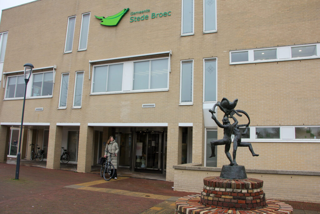 Voorgevel van het gemeentehuis van Stede Broec met een bronzen standbeeld op een bakstenen sokkel op de voorgrond.