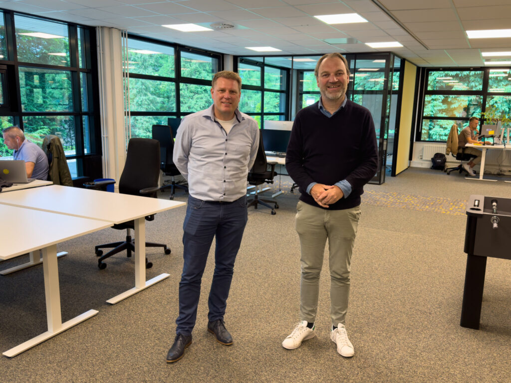 Twee mannen poseren lachend in een modern kantoor met werkplekken en grote ramen op de achtergrond.