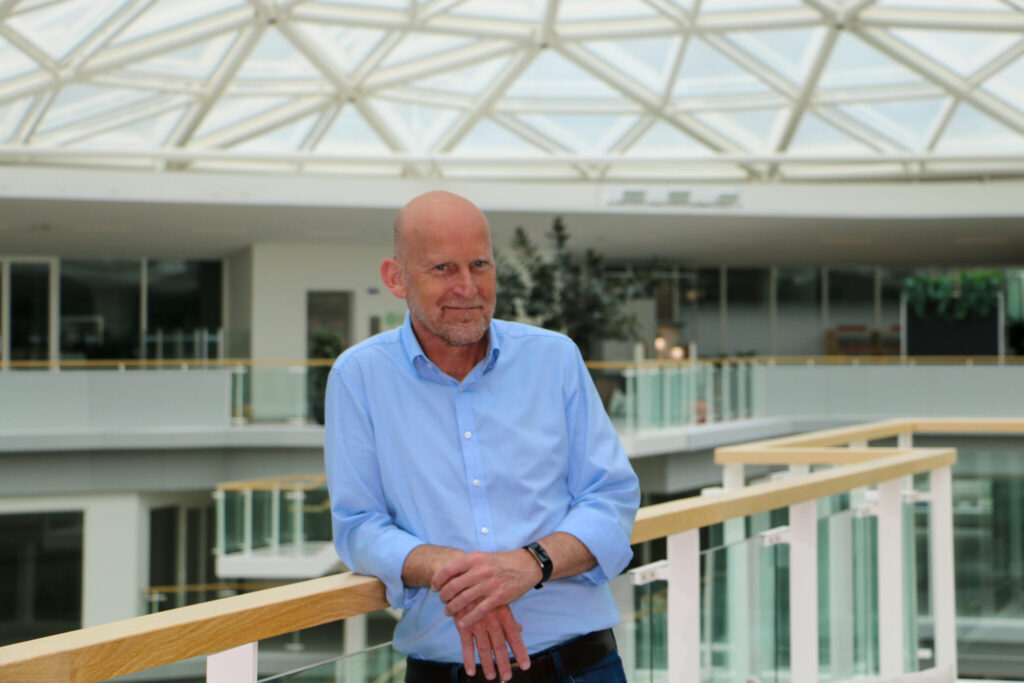 Man in een blauwe blouse leunt ontspannen op een balustrade in een modern, licht atrium met glazen dakconstructie.