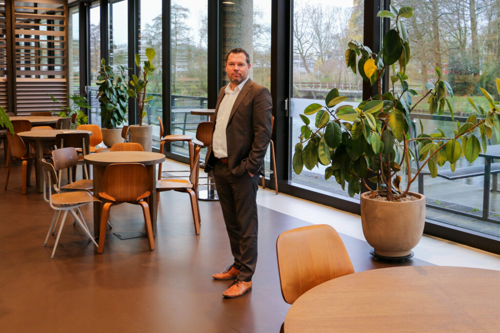 Man in pak staat in een moderne, lichte ruimte met houten tafels, stoelen en grote planten.
