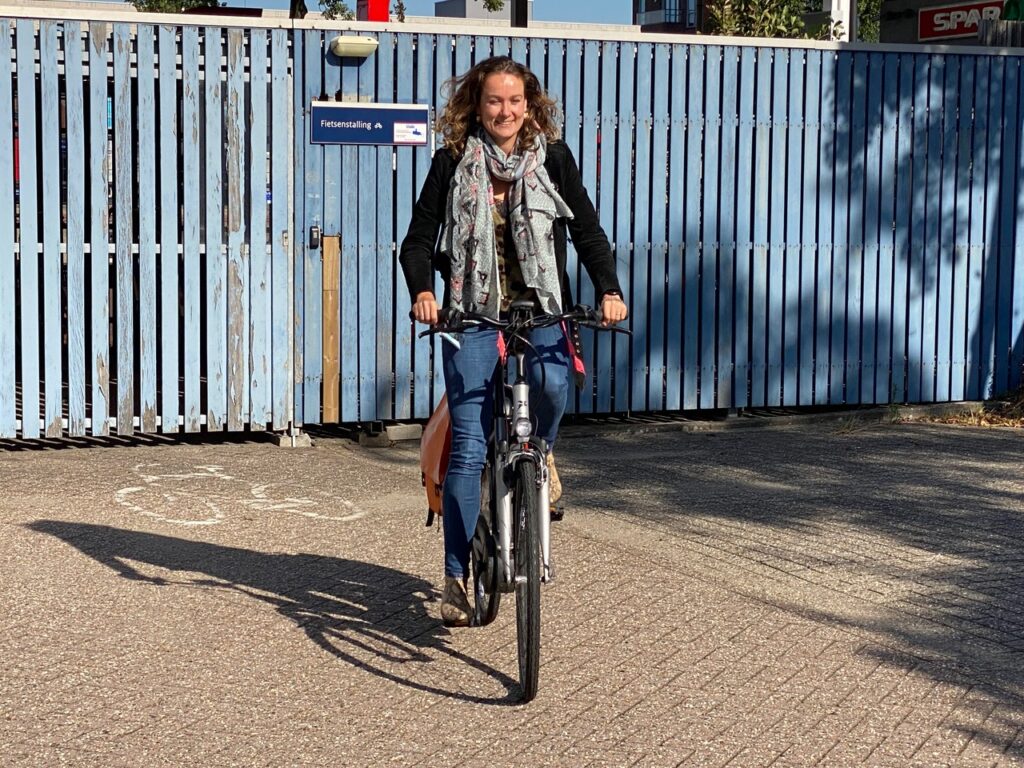 Vrouw fietst bij zonnig weer weg van een fietsenstalling met blauwe houten schutting.