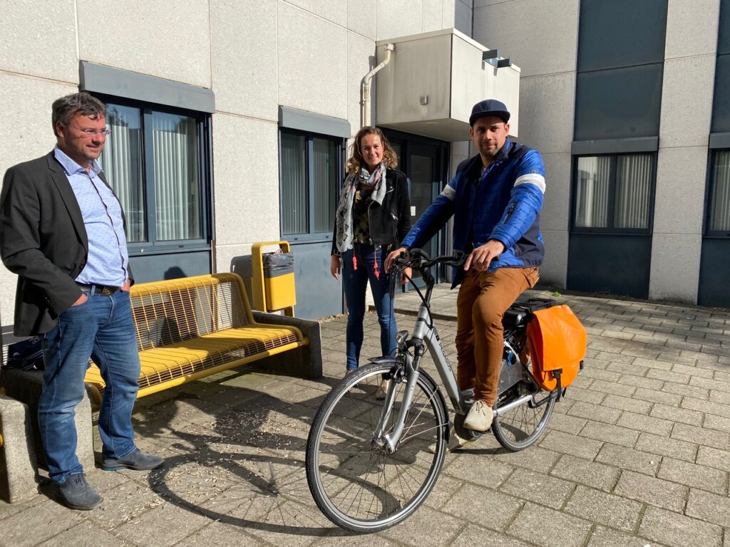Drie mensen staan buiten bij een gebouw, waarvan één man op een fiets zit met oranje fietstassen.