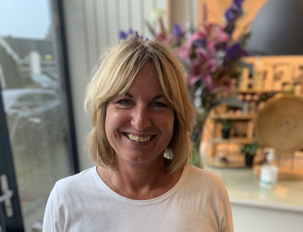 Vrouw met blond haar en witte trui lacht in een moderne, licht ingerichte ruimte met bloemen op de achtergrond.