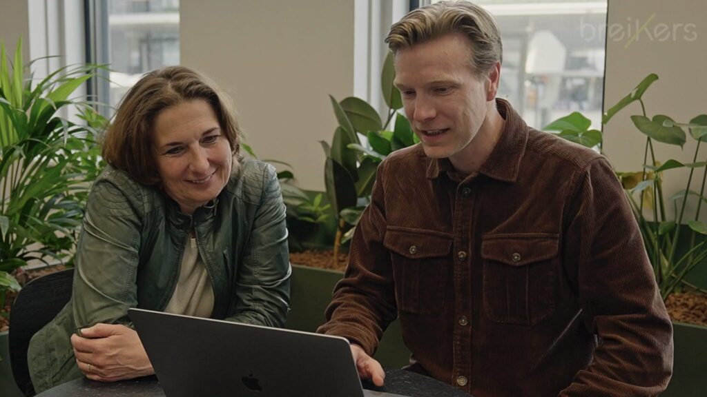 Twee mensen zitten samen aan een tafel en kijken glimlachend naar een laptop in een kantooromgeving met groene planten.