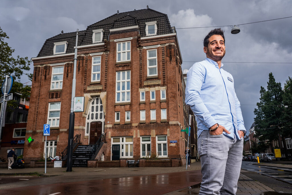 Man poseert lachend voor een groot, karakteristiek bakstenen gebouw onder een dreigende, bewolkte lucht.