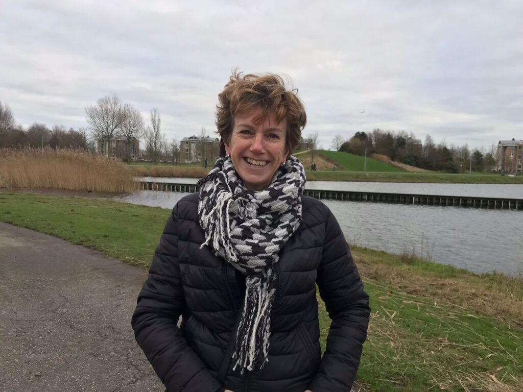 Vrouw met een zwart-witte sjaal en donkere jas staat glimlachend bij een waterpartij in een park, met op de achtergrond riet en gebouwen — Yos-Hopman.