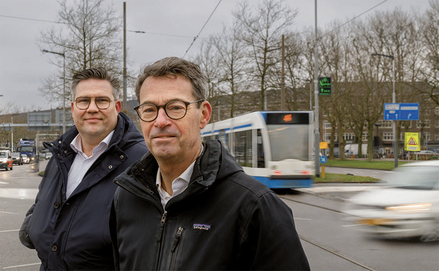 Twee mannen, waaronder Rein Aarts, poseren bij een druk kruispunt met voorbijrijdende tram en auto's.